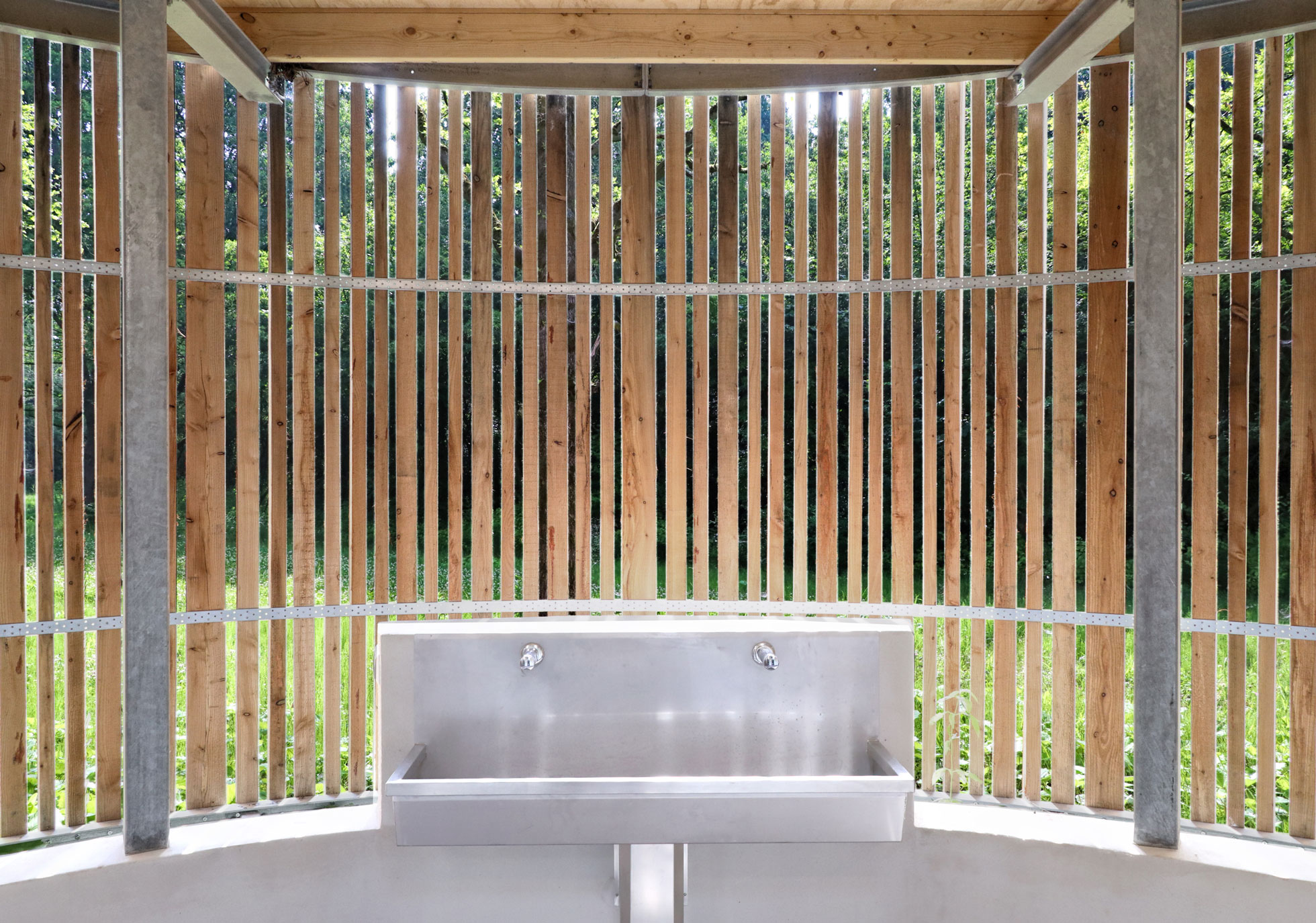 Interior view on the forest landscape through half open wooden façade, sunlight through wooden skin providing makes a sustainable and eco friendly solution. Metal sink offers view on green surrounding and air blows through for natural ventilation on the International Scout Centre in Zeewolde. The sanitary pavilions are designed and built by MOST Architecture,Rotterdam